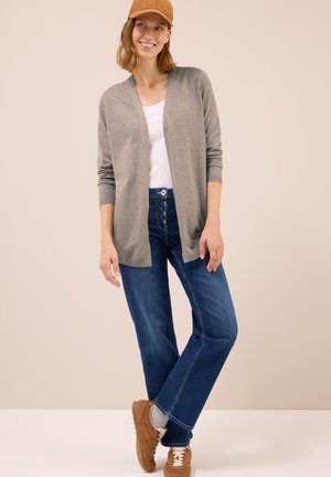 Lichtgrijze cardigan met open voorkant, gedragen over een wit shirt, gecombineerd met blauwe spijkerbroek en bruine sneakers, tegen een neutrale achtergrond.