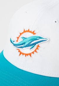 Hvit caps med teal flere som har en brodert delfinlogo omgitt av et oransje solstråledesign. Holdbart tekstil.