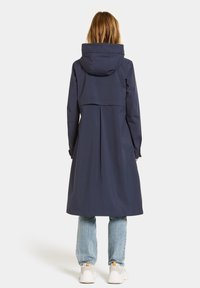 Manteau imperméable marine avec capuche, manches longues et coupe décontractée. Des pinces à l'arrière créent un léger détail plissé. Porté avec un jean bleu clair.