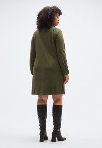 Olívazöld kordbársony ruha hosszú ujjal, térd alá érő szabással és oldalsó sliccekkel. Fekete, térdig érő, magas sarkú csizmával párosítva.