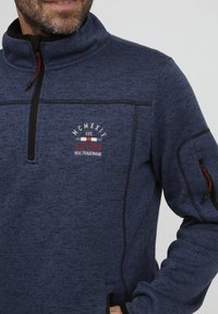 Jūrinės mėlynos spalvos fliso liemenė su aukštu apykakle, užtrauktuku priekyje ir raudonu akcentiniu siūlu. Turi krūtinės logotipą ir šonines kišenes su užtrauktuku.