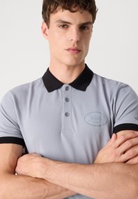 Lys grå polo skjorte med svart ribbestrikket krage og mansjetter, tre-knappers placket, brodert logo i mørk blå, glatt stofftekstur.
