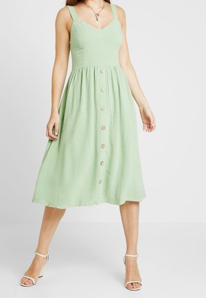 Vrouw die een mouwloze lichtgroene midi-jurk draagt met knopen aan de voorkant en beige enkellaarsjes met een blokhak, staand tegen een effen witte achtergrond.