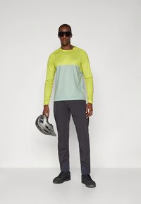 Majica z dolgimi rokavi z obliko barvnih blokov v svetlo zeleni in rumeni barvi, kombinirana s temno sivimi hlačami in črnimi supergami. Drži siv čelado.