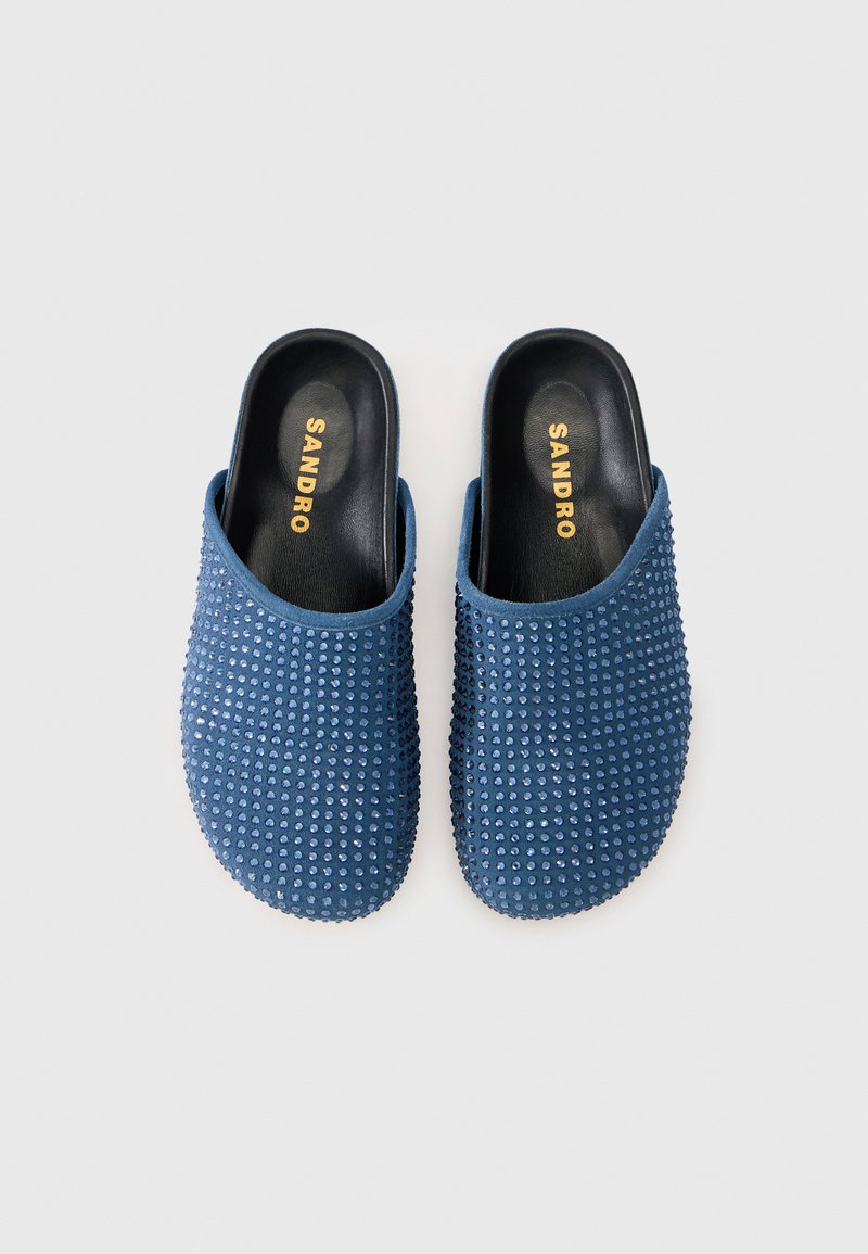 Blauwe instapmuilen met een gestructureerde bovenkant en kleine decoratieve studs. Zwarte binnenzolen met goudkleurige branding. Glad, afgerond silhouet.