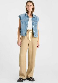 Denim vest met korte mouwen, lichtblauwe kleur, met voorzakken. Witte tanktop eronder. Losse beige linnen broek met trekkoord.