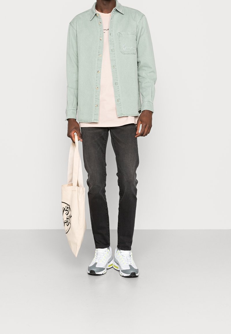 Homme portant une chemise vert clair boutonnée sur un t-shirt rose pâle, jean foncé, baskets blanches, tenant un sac fourre-tout crème avec impression noire.