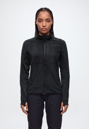 Une femme porte une veste polaire noire zippée, à col montant et poches zippées, sur un fond clair et uni.