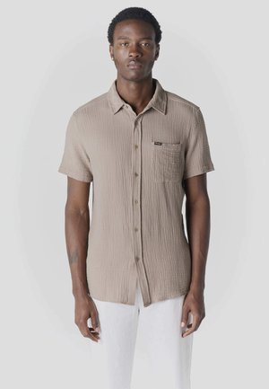 Hombre con camisa beige de manga corta abotonada con bolsillo en el pecho y pantalones blancos, de pie frente a un fondo liso gris claro.