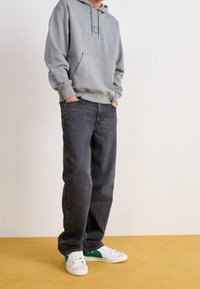 Felpa grigia con tasca frontale e cordino, abbinata a jeans scuri e sneakers bianche con dettagli verdi. Design e materiali semplici.
