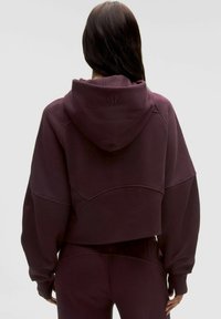 Kratka kapuca z dolgimi rokavi v temno burgundski barvi z mehko teksturo, širokimi rokavi in logotipom na hrbtu, v kombinaciji s usklajenimi hlačami.