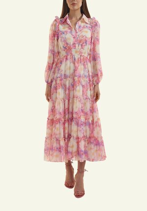 Frau trägt ein langes, gestuftes Blumenkleid in Pink-, Lila- und Cremetönen, mit aufgepufften langen Ärmeln und rosa Riemchen-Sandaletten mit hohen Absätzen.