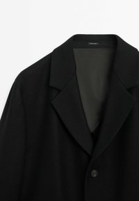 Manteau noir à simple bouton avec revers à encoche et doublure intérieure lisse, étiqueté "Fabriqué en Égypte", présenté sur un fond blanc.