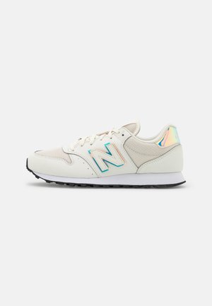 Hvid og lys beige New Balance-sneaker med mesh-paneler, snørelukning foran, iriserende hælflap og sort rillede sål, vist fra siden.