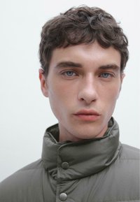 Kapuzen-Puffermantel in Olivgrün, mit Druckknopfverschluss, glatter Textur und minimalistischem Design. Die Umgebung ist neutral und schlicht.