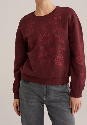 Personne portant un sweat-shirt grenat avec une broderie florale rouge, associé à un jean en denim gris, debout devant un fond uni.