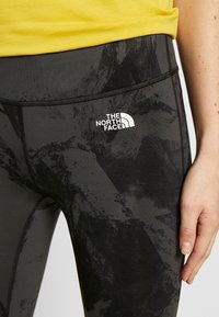 Czarne legginsy z wysokim stanem, z teksturowanym wzorem w różnych odcieniach szarości, z białym logo na górnej części uda.