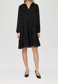 Černé šaty s volány, dlouhými rukávy a výstřihem do V, zdobené vertikálními pruhy a vzdušnou siluetou, vyrobené z lehkého, průsvitného materiálu.