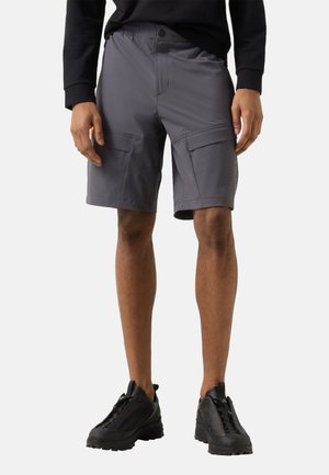 Mężczyzna w szarych szortach typu cargo i czarnych trampkach, stoi z rękami swobodnie opuszczonymi wzdłuż ciała na prostym jasnym tle.