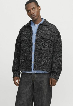 Black and white speckled jacket with two chest pockets, button closure, and a relaxed fit, worn over a light denim shirt.