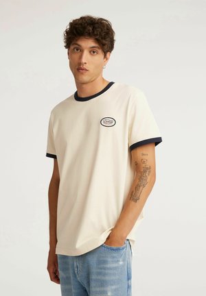 Młody mężczyzna z kręconymi włosami, ubrany w kremowy t-shirt z czarnym obszyciem i niebieskie dżinsy, pokazujący wytatuowane lewe przedramię.