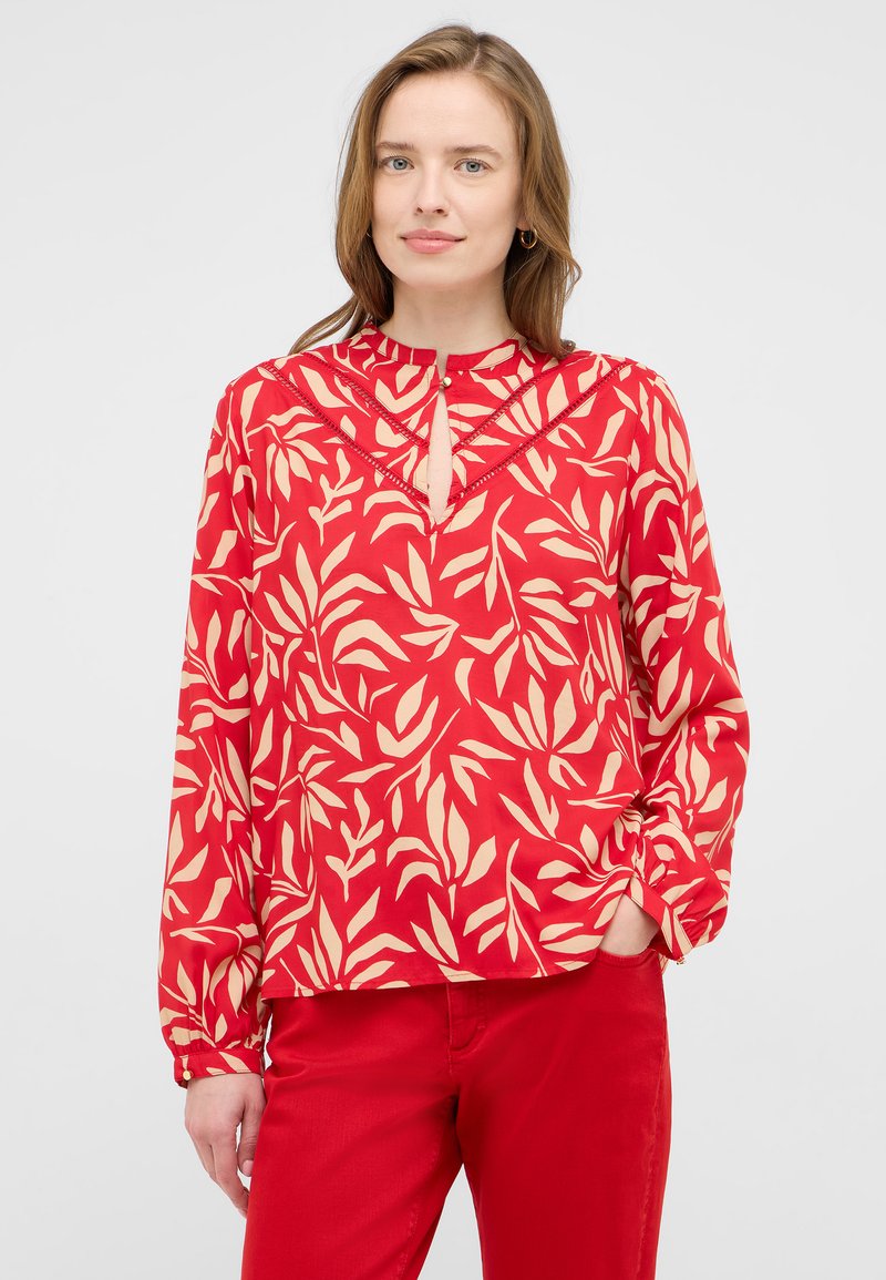 Blouse rouge avec un motif floral crème, manches longues, encolure en forme de goutte et détails en dentelle. Fabriquée dans un matériau léger et lisse.