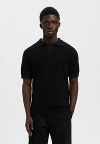 Ung man som bär en svart kortärmad polo och svarta byxor, står mot en helt vit bakgrund.