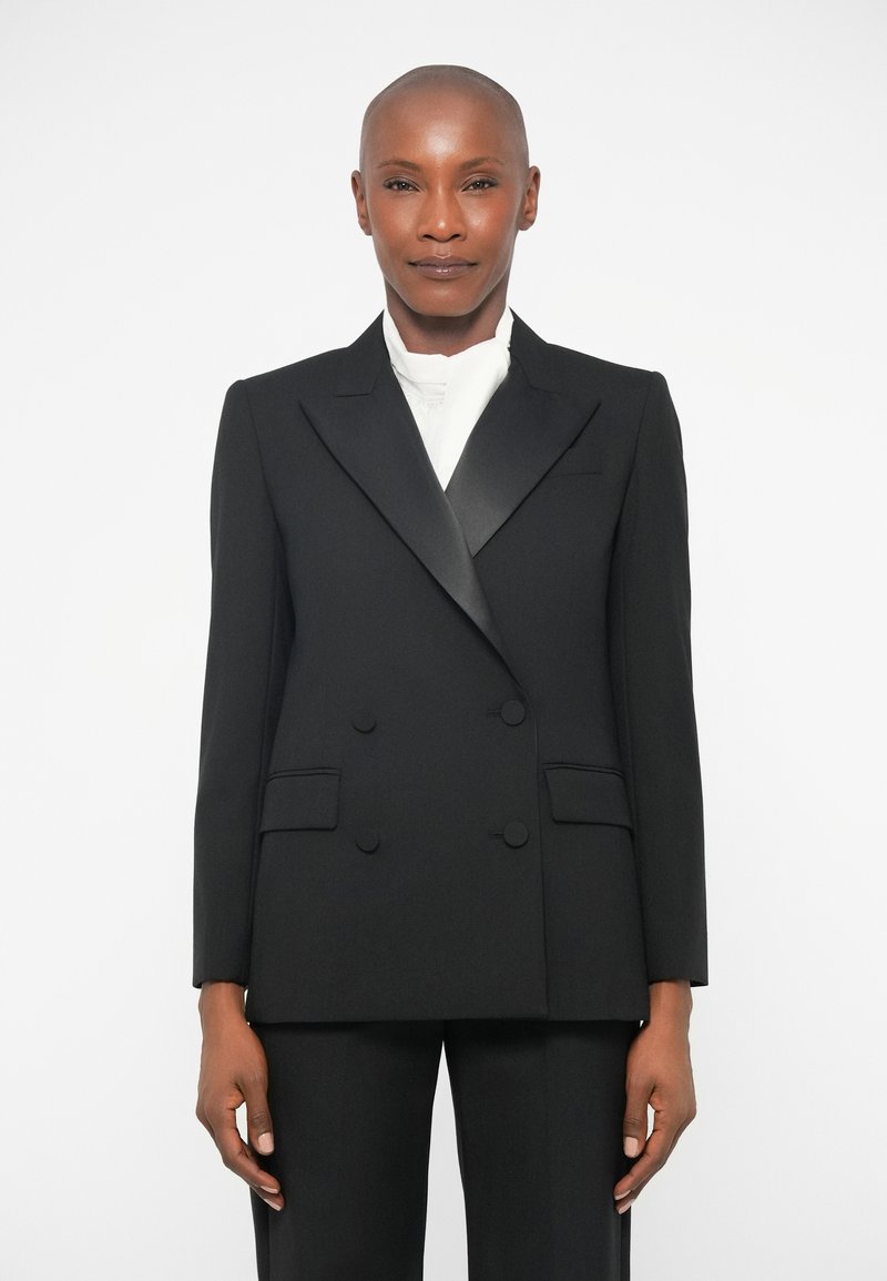 Blazer noir à double boutonnage avec revers crantés, en tissu lisse. Il dispose de deux poches avant et d'un pantalon noir assorti.