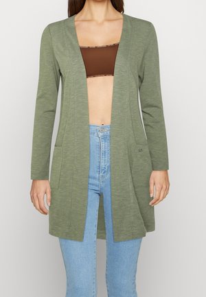 Žena má na sobě otevřený olivově zelený dlouhý svetr s kapsami, hnědý krátký top a světle modré džíny na šedém pozadí.