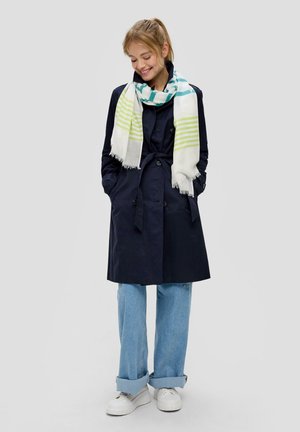 Marineblauwe trenchcoat met knopen aan de voorkant, zijzakken en een strikriem, gecombineerd met lichtblauwe wijde jeans en een gestreepte wit-groene sjaal.