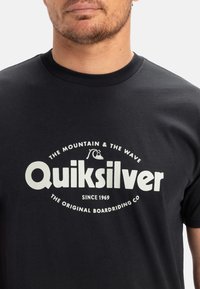 Černé bavlněné tričko s bílým kruhovým logem a textem. Hladký crew výstřih, krátké rukávy, jednoduchý design a jemná textura.