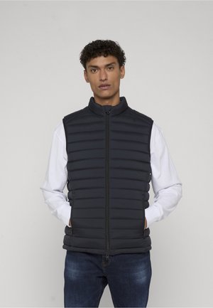 Mand iført sort quiltet ærmeløs vest over hvid langærmet skjorte og blå jeans, stående med hænderne i vestens lommer.