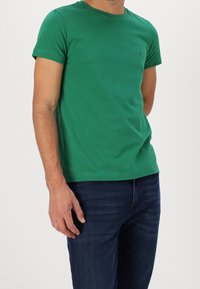 Grünes Baumwoll-T-Shirt mit Rundhalsausschnitt, kurzen Ärmeln und einem kleinen Logodetail auf der Brust. Kombiniert mit dunkelblauen Jeans.