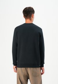 Black sweatshirt featuring long sleeves and a crewneck, made from soft fabric. Visible stitching on cuffs, plain back, paired with beige pants.