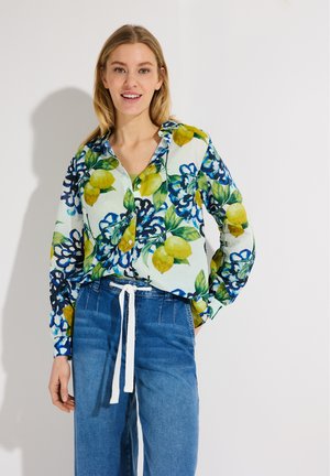 Lächelnde Frau trägt ein buntes Hemd mit Zitronen- und Blumenmustern, kombiniert mit hochgeschnittenen blauen Jeans, die mit einem weißen Kordelzug gebunden sind.