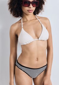 Hvit teksturert bikinioverdel med slanke stropper, kombinert med svart og hvit stripete bikinitruser. Modellen bruker store røde solbriller.