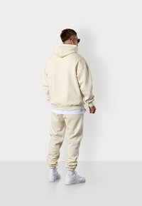 Haut à capuche crème avec cordon de serrage et poignets côtelés, associé à un jogging crème assorti avec des rayures sur les côtés. Des baskets blanches sont portées.