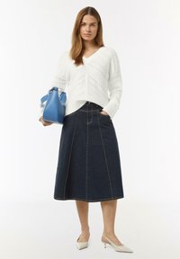 Witte gebreide v-hals trui met open weefsel, donkere spijkerrok in A-lijn met contrasterende stiksels, en lichtgekleurde slingback hakken, met een blauwe handtas.