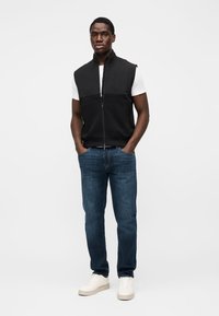 Μαύρο γιλέκο με φερμουάρ και ψηλό γιακά πάνω από λευκό t-shirt, συνδυασμένο με μπλε τζιν και λευκά αθλητικά παπούτσια. Απλός σχεδιασμός, καθημερινή εμφάνιση.