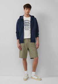 Marineblauem Hoodie mit Kordelzügen über einem weißen Grafik-T-Shirt, kombiniert mit khakifarbenen Shorts und hellen Turnschuhen mit beigen Akzenten.