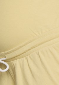 Tecido amarelo claro com uma textura suave, apresentando uma cintura elástica com cordão branco e detalhe franzido na borda inferior.