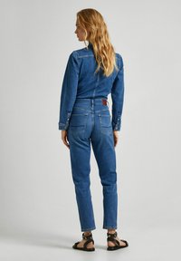 Džínový overal ve středně modré barvě, přiléhavý střih s dlouhými rukávy, zapínáním na knoflíky na manžetách a přiléhavým pasem. Obsahuje zadní kapsy a koženou nášivku.