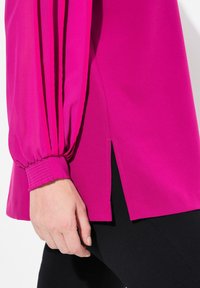 Blusa fucsia a maniche lunghe con tessuto plissettato e un'apertura laterale, caratterizzata da polsini elasticizzati e una superficie liscia. Indossata sopra pantaloni neri.