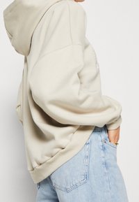 Beige Kapuzenpullover mit lockerer Passform, ausgestattet mit einer Kordelzugkapuze und gerippten Bündchen, kombiniert mit hellblauen Jeans. Glatte Stofftextur.
