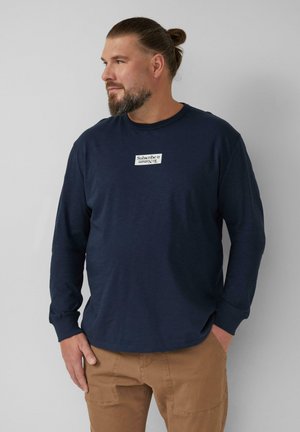 Marineblauwe longsleeve shirt van lichtgewicht stof met een ronde hals en een logo-patch met tekst aan de voorkant. Gecombineerd met tan kleurige cargobroek.
