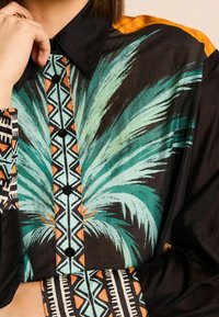 Chemise boutonnée arborant des imprimés de feuilles de palmier vibrants en bleu sarcelle et orange, avec des motifs géométriques en noir et blanc le long du col et des manches.