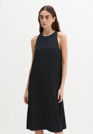 Frau mit langen dunklen Haaren, die ein ärmelloses, locker sitzendes, knielanges schwarzes Kleid vor einem schlichten weißen Hintergrund trägt.