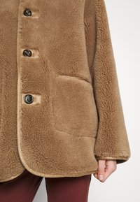 Brun, knappet, tekstureret fleecetrøje med stor forlomme og lange ærmer, båret over bordeauxbukser, hånd synlig ved siden.