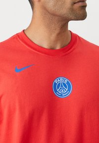 Rød bomuld t-shirt med et blåt Nike-logo på venstre skulder og et cirkulært blåt Paris Saint-Germain-våbenskjold i midten.