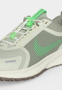 Ανοιχτό γκρι και πράσινο αθλητικό παπούτσι Nike με διχτυωτό άνω μέρος, λογότυπο με πράσινες ρίγες, λευκή σόλα και πράσινα κλειδώματα κορδονιών.